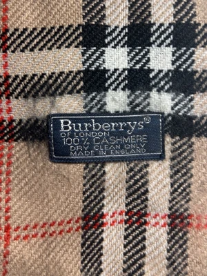 Beige rutig halsduk från Burberrys - Klassisk beige halsduk från Burberrys i 100% kashmir med ikoniskt rutmönster i svart, vitt och rött. Halsduken har fransar i ändarna och är tillverkad i England. Perfekt accessoar för att lyfta din outfit.