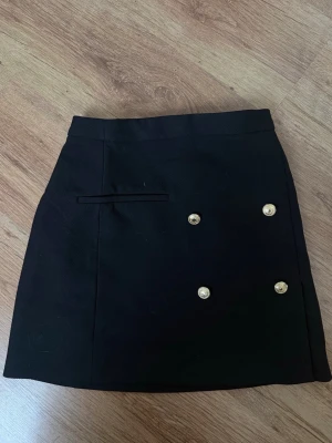 Svart kortkjol med guldknappar - Stilren svart kortkjol med fyra dekorativa guldknappar framtill och en diskret ”fakeficka”. Kjolen har dragkedja i sidan för enkel på- och avtagning. Perfekt för dig som gillar klassisk och chic stil.