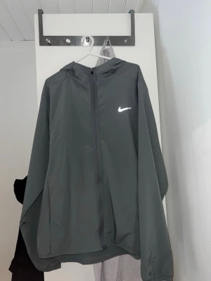 Mörkgrå Nike vindjacka med huva - Snygg mörkgrå vindjacka från Nike med hel dragkedja framtill och huva. Jackan har en minimalistisk design med den klassiska vita Nike-loggan på bröstet. Tillverkad i lätt och vindtätt material, perfekt för blåsiga dagar.