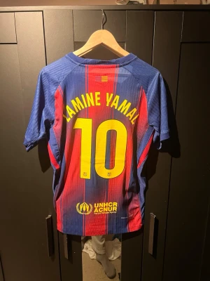 FC Barcelona matchtröja Lamine Yamal - Säljer en officiell FC Barcelona matchtröja från Nike med Lamine Yamal och nummer 10 på ryggen. Tröjan är blå och röd med gula detaljer, klubbmärke och sponsorer på bröstet. Tillverkad i lätt och ventilerande material, perfekt för fotbollsträning eller match.