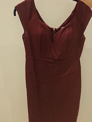 Vinröd kort klänning med dragkedja - Snygg vinröd kort klänning med markerad midja och djup v-ringning. Klänningen har en dekorativ dragkedja i ryggen och är tillverkad i ett slätt, stretchigt material. Perfekt för dig som vill ha en elegant och stilren look.