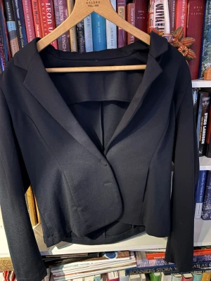 Svart kort kavaj med enkelknäppning - Stilren svart kavaj med enkelknäppning och klassisk krage. Modellen är kortare och har figurnära passform med långa ärmar. Perfekt för en trendig och clean look. Materialet ser ut att vara en slät, vävd.