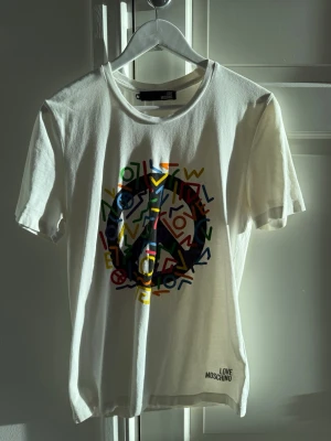 Vit t-shirt från Love Moschino - Säljer en vit t-shirt från Love Moschino med ett stort färgglatt peace-märke i grafiskt mönster på bröstet. T-shirten har rund hals och korta ärmar. Märkeslogga tryckt nere vid fållen. Perfekt för dig som gillar statement-plagg!