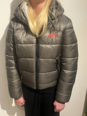 Grå pufferjacka från Superdry - Snygg grå pufferjacka från Superdry med huva och dragkedja framtill. Jackan har två fickor med dragkedja och logga i rött på bröstet. Perfekt för kalla dagar och ger en cool, sportig look. Jackan är 38 men mer som en S