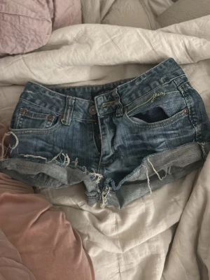 Blå jeansshorts med fransar - Säljer ett par blå jeansshorts från Jeane med råa kanter och fransar. Shortsen har klassisk femficksdesign, knappgylf och uppvikta benslut. Perfekta för varma dagar och en avslappnad stil. Vet inte storlek men tror xs