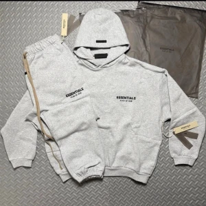  Essentials Fear of God - Snygg ljusgrå hoodie från Essentials Fear of God med svart logga tryckt på bröstet. Klassisk oversized passform, ribbade muddar och stor huva. Tillverkad i mjuk bomullsblandning som känns riktigt skön mot huden. Perfekt för dig som gillar streetwear och enkel stil.