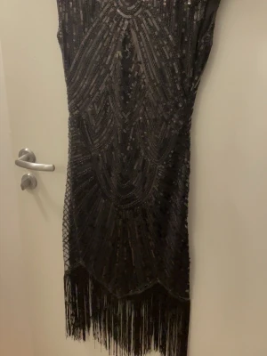 Svart glittrig fransklänning - Säljer en svart klänning med glittriga paljetter och dekorativa mönster. Klänningen har fransar längs nederkanten och är figurnära med ärmlös design. Perfekt för dig som vill sticka ut med en elegant och unik look.