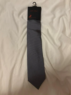 Mörkgrå slips i 100% siden - Säljer en stilren mörkgrå slips i 100% siden med diskret rutmönster. Perfekt accessoar för att lyfta din outfit med en klassisk touch. Slipsen har en smal spets och elegant finish.