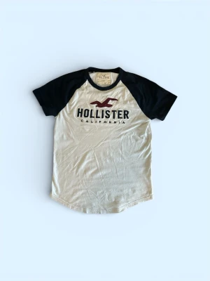  Hollister tröja - Snygg t-shirt från Hollister med svart-vita kontraster och klassisk logga på bröstet. T-shirten har svarta korta ärmar och vit kropp, samt rund halsringning. Perfekt till jeans för en avslappnad och trendig look.