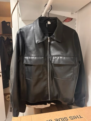 Svart skinnjacka från H&M - Snygg svart skinnjacka från H&M med rak passform och två stora bröstfickor med lock. Jackan har klassisk krage och dragkedja framtill. 
