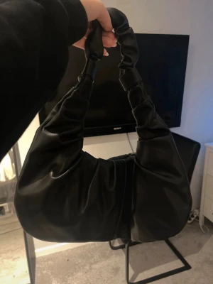 Svart axelväska i fejkläder - Trendig svart axelväska från NA-KD i mjukt fejkläder. Väskan har en cool, rynkad design och ett brett handtag som ger en modern vibe. Perfekt storlek för det viktigaste och enkel att matcha med olika outfits. Köpt second hand men kommer tyvärr inte till användning! Skriv vid frågor!