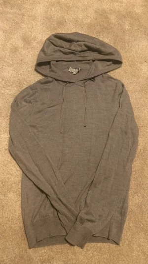 Grå hoodie från All Good Clothing - Säljer en stilren grå hoodie från AHLQUIST Clothing i storlek XXS. Hoodien har dragsko i huvan och är tillverkad i ett tunt, mjukt merinoull som känns skönt mot huden. Perfekt för dig som gillar en enkel och clean look.