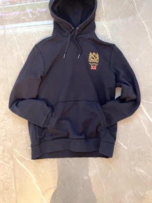 Svart hoodie från Morris - Snygg svart hoodie från Morris med broderad logga och brittisk flagga på bröstet. Klassisk känguruficka framtill och justerbar dragsko i huvan. Perfekt för en chill och stilren look. Tillverkad i mjukt material som känns skönt mot huden.