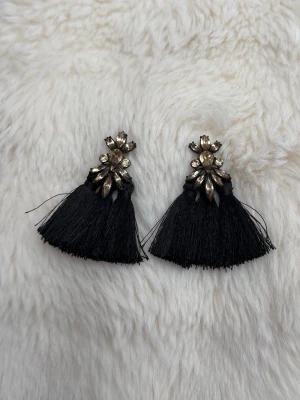 Svarta tofsörhängen med strass - Unika örhängen med svarta tofsar och gnistrande strasstenar i guldtonad metall upptill. Örhängena har en dekorativ, blomliknande form och ger en cool statement-look. Perfekta för dig som vill sticka ut med accessoarerna. Köpta i Italien 