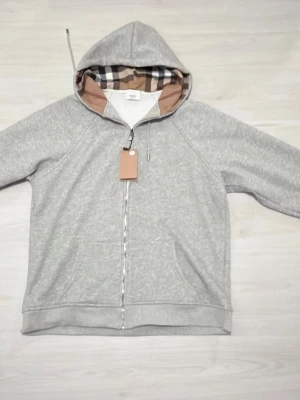 Grå zip hoodie från Burberry - Snygg grå zip hoodie från Burberry med klassiskt rutigt foder i huvan. Hoodien har dragkedja framtill, dragsko i huvan och diskret broderad logga på bröstet. Perfekt för en avslappnad men stilren look. Tillverkad i mjukt bomullsmaterial.