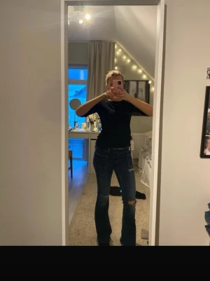 Mörkblå slitna jeans med hål - Säljer ett par mörkblå jeans med tight passform och slitna detaljer, inklusive hål på knäna. Jeansen har klassisk femficksdesign och är i stretchigt denimtyg. Perfekta för en trendig och avslappnad stil. Virala jeans från Gina