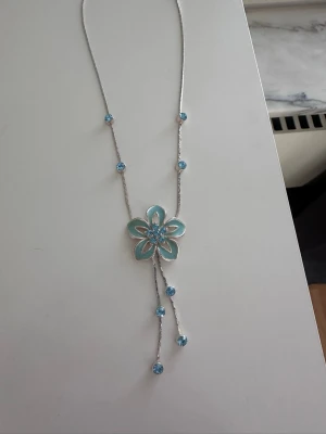 Silverhalsband med blå blomma - Snyggt halsband i silverfärg med en stor blå blomma som hänge. Halsbandet har små blå pärlor längs kedjan och två hängande kedjor med pärlor nedanför blomman. Köpt second hand, mycket bra skick!