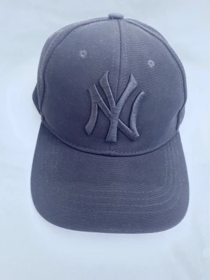 Mörkgrå New York Yankees keps - Snygg mörkgrå keps från New Era med klassisk New York Yankees-logga broderad framtill i samma färg som kepsen. Kepsen har böjd skärm och ventilerande hål upptill. Tillverkad i slitstark bomull, perfekt för en sportig och avslappnad stil.  Jag samlar på kepsar, så de är sparsamt använda och alla har kvar stuket från när de var nya. När jag skickar en keps så gör jag det med mycket vaddering och alltid i en hård kartong. ( är nog ensam om för begagnade kepsar. 