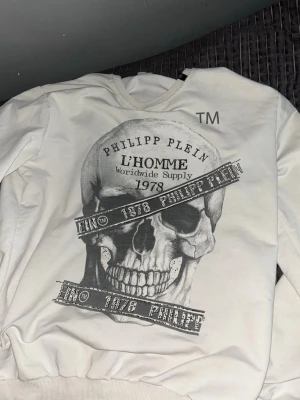 Vit sweatshirt från Philipp Plein - Säljer en vit sweatshirt från Philipp Plein med stor döskalle- och textprint på framsidan. Tröjan har rund halsringning, ribbade muddar och en liten logotypetikett i sidan. Materialet känns mjukt och bekvämt, perfekt för en edgy streetwear-look.