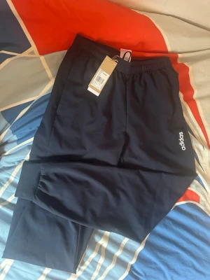 Marinblå adidas mjukisbyxor S - Snygga marinblå mjukisbyxor från adidas med vit logga på benet. Byxorna har resår i midjan, muddar vid sopparna och raka ben. Perfekta för träning eller chill. Tillverkade i ett mjukt och bekvämt material.