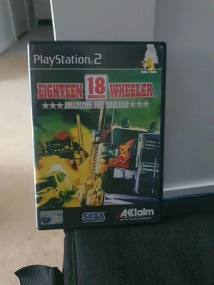 Eighteen Wheeler till PS2 - Ett arkadigt racingspel till PlayStation 2 där du kör massiva lastbilar genom USA. Omslaget är färgstarkt med en stor lastbil i fokus och explosioner i bakgrunden. Spelet är utvecklat av SEGA och utgivet av Acclaim. Perfekt för dig som gillar fartfyllda truckrace.