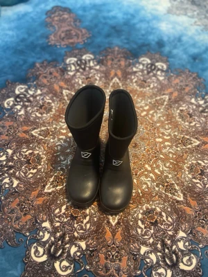 Svarta gummistövlar från Didriksons - Säljer ett par svarta gummistövlar från Didriksons med högt skaft och logga på sidan. Stövlarna är tillverkade i slitstarkt gummimaterial och har en klassisk, stilren design som passar perfekt för regniga dagar.