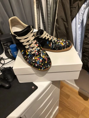Mason margiellas med färg - Unika svarta sneakers från Maison Margiela med färgstänk i blått, gult, vitt och rött. Skorna har beige snören och klassisk gummisula. Materialet är mocka och läder, och designen är inspirerad av German Army Trainer. Perfekt för dig som vill sticka ut med din stil. Hör av om frågor.