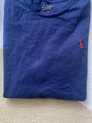 Marinblå t-shirt från Polo Ralph Lauren - Klassisk marinblå t-shirt från Polo Ralph Lauren med den ikoniska röda logotypen broderad på bröstet. Tillverkad i mjuk bomull och har en normal passform. Perfekt basplagg för en clean och stilren look.