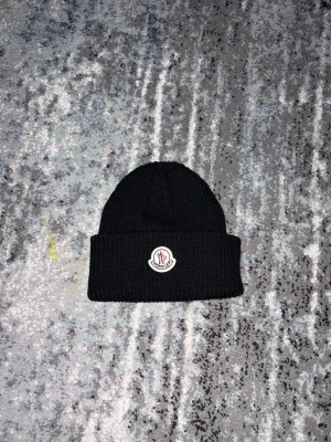 Svart ribbstickad mössa från Moncler går och sänka priset  - Säljer en svart ribbstickad mössa från Moncler med uppvikt kant och Moncler-logga framtill. Klassisk modell i mjukt material som passar perfekt till kalla dagar. Enkel och stilren design som funkar till det mesta.