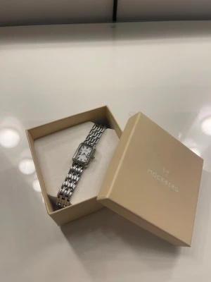 Mockberg silverfärgad klocka - Stilren klocka från Mockberg med rektangulär vit urtavla och silverfärgat armband i metall. Klassisk design med länkarmband och tydliga svarta siffror på urtavlan. Perfekt accessoar för dig som gillar minimalistisk och elegant stil.
