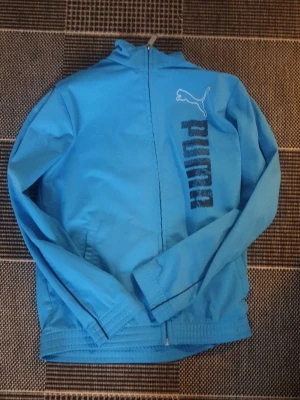 Blå vindjacka från Puma - Snygg blå vindjacka från Puma med dragkedja framtill och logga tryckt på bröstet. Jackan har hög krage, långa ärmar och ribbade muddar vid ärmslut och nederkant. Perfekt för en sportig look.