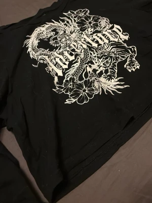 Svart croppad topp med draktryck - Cool svart croppad topp med vitt drak- och blommönster framtill. Långärmad modell med edgy streetstil, perfekt för dig som gillar statement-plagg. Trycket är detaljerat och täcker större delen av framsidan.