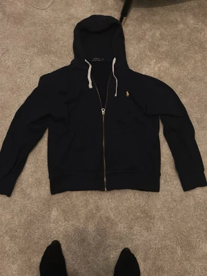 Mörkblå hoodie från Polo Ralph Lauren - Snygg mörkblå hoodie från Polo Ralph Lauren med dragkedja framtill och gul logga broderad på bröstet. Tröjan har huva med vita snören och två fickor fram. Tillverkad i mjukt bomullsmaterial och har ribbade muddar vid ärmslut och nederkant.