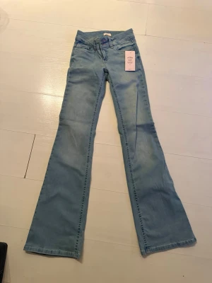 Ljusblå bootcut jeans från Wrangler - Säljer ett par ljusblå bootcut jeans från Nelly med klassisk femficksdesign och rosa broderad detalj på bakfickorna. Jeansen har låg midja och utsvängda ben, perfekta för en retroinspirerad look. Materialet är mjuk denim i bomull.