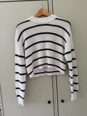 Vit stickad tröja med svarta ränder - Säljer en vit stickad tröja med svarta horisontella ränder. Tröjan har en avslappnad passform, ribbade muddar och är lite croppad i modellen. Perfekt för dig som gillar en enkel men trendig look.
