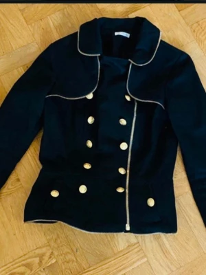 Ida Sjöstedt Svart kavaj med guldknappar - Stilren svart kavaj med dubbelknäppning och dekorativa guldknappar framtill. Kavajen har en unik krage med kontrasterande kantband och markerad midja med volang baktill. Perfekt för dig som vill sticka ut med en elegant och trendig look. Väldigt fint skick.