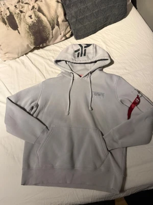 Alpha Industries hoodie - Snygg ljusgrå hoodie från Alpha Industries med svart logga på huvan och bröstet. Har klassisk känguruficka, dragsko i huvan och en röd tagg på ärmen. Perfekt för en avslappnad streetwear-look. Använd upp till 3-4 gånger. 10/10 skick, inga skador, slitningar alls. 