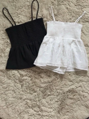 Svart och vit smockad linnetopp - Två söta linnetoppar i svart och vitt med smockad överdel och tunna axelband. Topparna har en volangig nederdel och är perfekta för varma dagar. Tillverkade i mjukt bomullstyg som känns skönt mot huden. (Den svarta toppen är ifrån Gina Tricot i storlek XS, den vita toppen är ifrån Gina Young i storlek 158/164.)