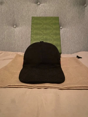 Svart keps från Gucci med röd/grön rand - Säljer en svart keps från Gucci med klassisk böjd skärm och diskret GG-mönster i tyget. På sidan finns den ikoniska röd-gröna randen. Justerbar rem baktill för perfekt passform. Perfekt accessoar för dig som vill ha en stilren och lyxig look. ÄKTA! Kvitto på sista bilden 