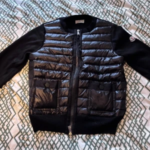 Svart dunväst från Moncler - Snygg svart dunväst från Moncler med quiltad framsida och två stora fickor med knappstängning. Ärmar och rygg i mjukt tyg, rund halsringning och dragkedja framtill. Moncler-logga på ärmen för extra stilpoäng. (KVITTO FINNS)