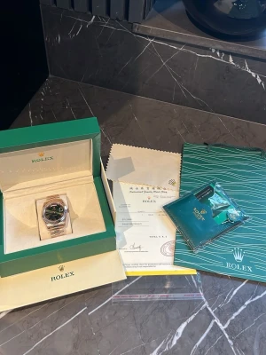 Rolex klocka med grön urtavla - Lyxig Rolex klocka med grön urtavla och armband i guldtonad metall. Klassisk rund form och elegant design, levereras i originalbox med certifikat och tillbehör. Perfekt för dig som vill ha något exklusivt och tidlöst.