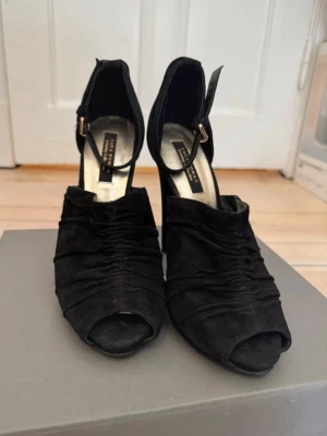 Svarta peep toe pumps från Zara - Snygga svarta pumps från Zara med peep toe och hög klack. Skorna har rynkade detaljer i mockaimitation och justerbar ankelrem med guldfärgat spänne. Perfekta för dig som vill ha en elegant och trendig look.