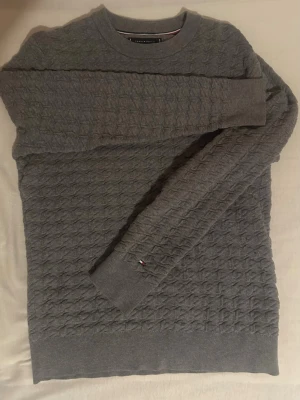 Grå stickad tröja Tommy Hilfiger - Säljer en grå stickad tröja från Tommy Hilfiger med diskret strukturmönster över hela plagget. Tröjan har rund halsringning och långa ärmar. Liten broderad logga på ärmslutet ger en snygg detalj. Perfekt till höst och vinter!