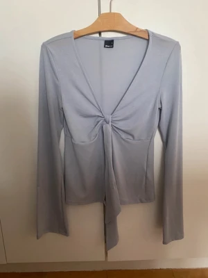 Ljusgrå långärmad topp med knyt - Trendig ljusgrå långärmad topp från Gina Tricot med v-ringning och snygg knytdetalj framtill. Tillverkad i mjuk och stretchig polyesterblandning som sitter skönt på kroppen. Perfekt för dig som gillar stilrena och moderna plagg. Endast använd en gång, men köpt på vinted sedan innan. Lite nopprig under armarna men annars tillkommer inga defekter eller skavanker. 