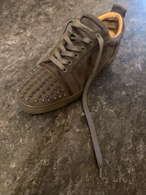 Gråa Christian Louboutin sneakers - Unika grå sneakers från Christian Louboutin med nitar på tån och klassisk låg siluett. Skorna har grå snörning, mjuk insida i beige och är tillverkade i mocka. Perfekta för dig som vill sticka ut med en edgy och lyxig stil.