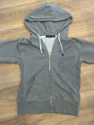 Grå zip hoodie från Polo Ralph Lauren - Grå hoodie med dragkedja från Polo Ralph Lauren. Tröjan har huva med vita snören. Storleken är XXS och har passat kille 158 cm.