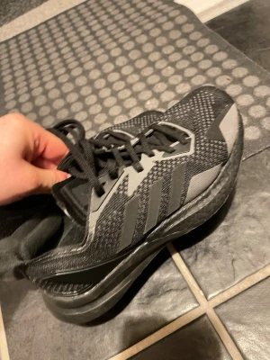 NYA SVARTA Adidas sneakers - Snygga svarta sneakers från Adidas med grå ränder på sidorna och mönstrad ovandel i mesh. Skorna har klassisk snörning och en tjock, bekväm sula som ger bra stöd. Perfekta för dig som gillar sportig stil och vill ha något som funkar till det mesta.