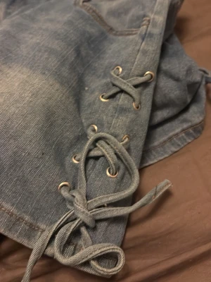 Blå jeansshorts med snörning - Snygga blå jeansshorts med cool snörningsdetalj i sidorna och klassiska bakfickor. Perfekta för varma dagar och ger en trendig touch till din outfit. Gjorda i slitstarkt denimtyg.