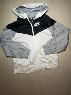 Nike vindjacka med huva svart/grå/vit - Nike vindjacka i färgerna svart, grå och vit. Jackan har huva, hel dragkedja framtill och två sidofickor med dragkedja. Stort Nike-logotyp tryckt på bröstet. Tillverkad i lätt och vindtätt material, perfekt för blåsiga dagar.Windbreaker! Pris kan diskuteras!😀