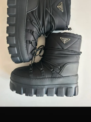 Svarta snowboots - Stilrena svarta moon boots från Panda med chunky sula och snörning runt hela skaftet. Ovandelen är i slitstark textil och insidan är fodrad för extra värme. Bootsens höga sula ger grymt lyft och edgy look. Perfekta för kalla dagar när du vill hålla stilen. Står storlek 39 men är som 38, insidan sitter löst och är uttagbara.